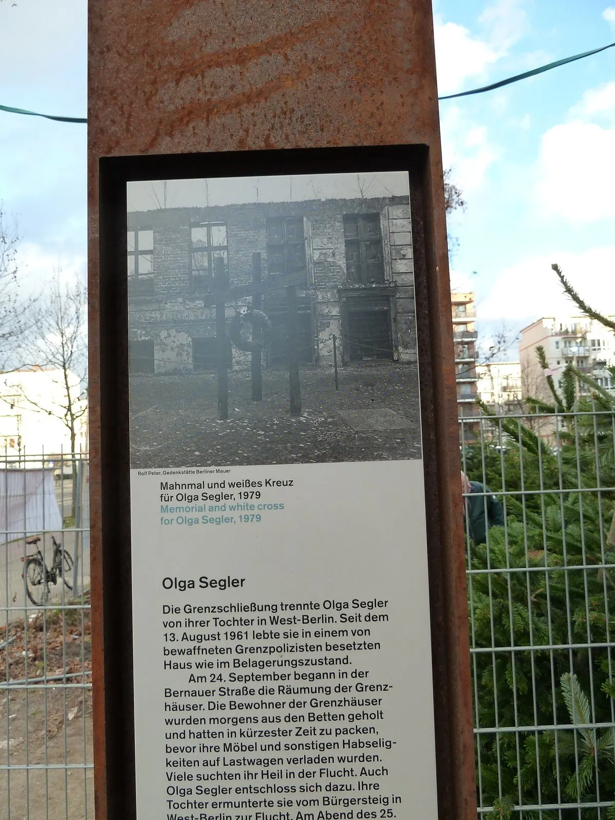Rusted steel stele with a historical photo of a memorial and white cross and text headed "Olga Segler"