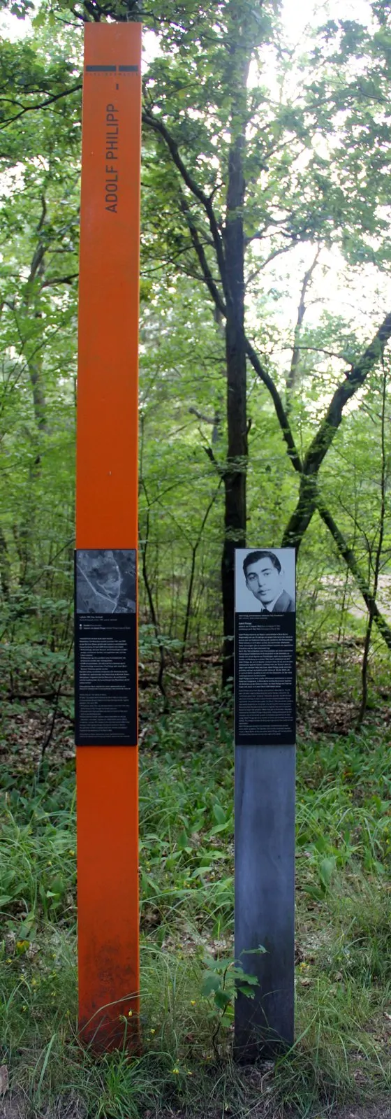 Tall orange column inscribed 'Adolf Philipp' and a grey post with his portrait panel, in green woodland