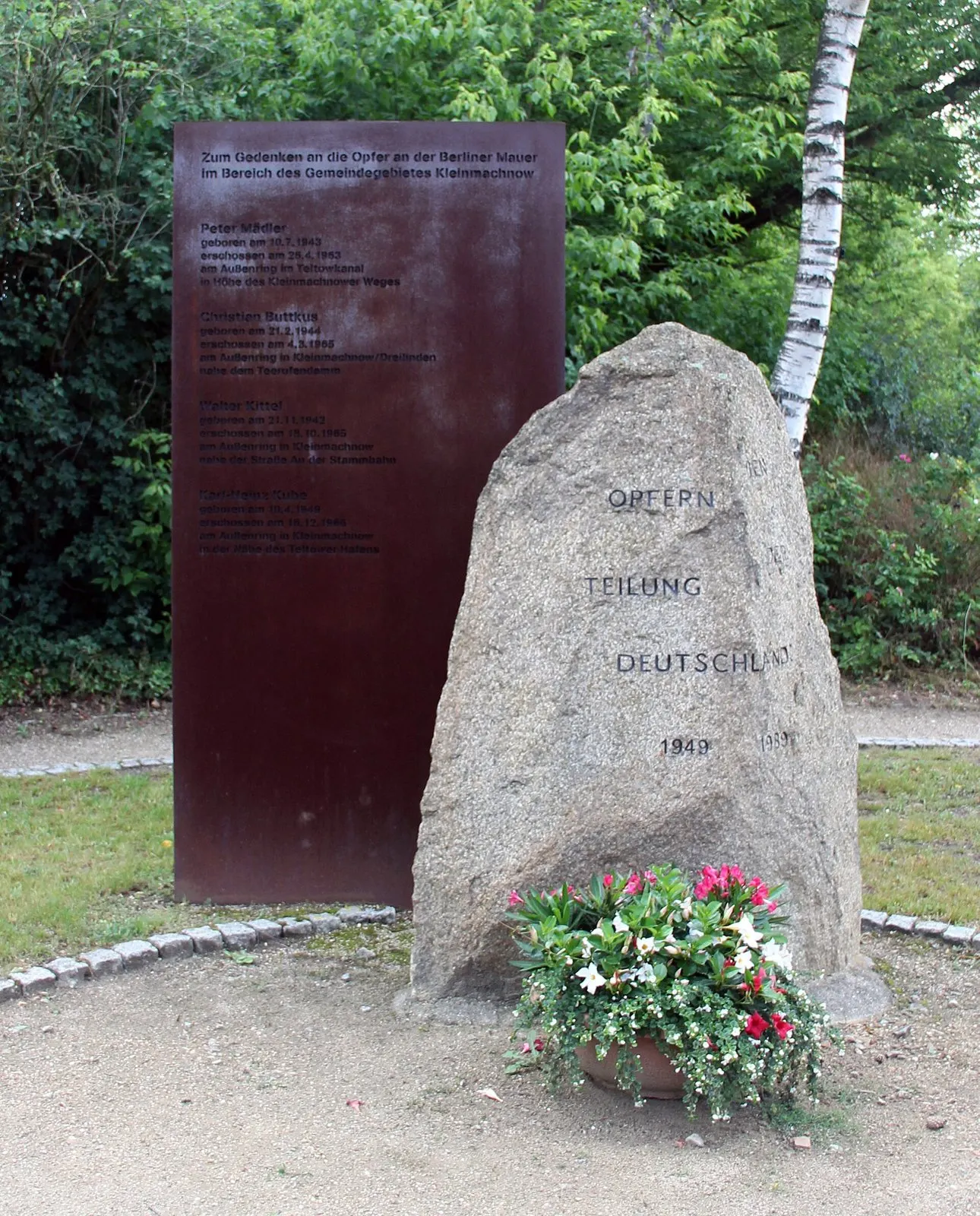 Rust-coloured steel plaque with a list of names beside a granite boulder inscribed 'Opfern Teilung Deutschlands 1949', with a pot of red and white flowers