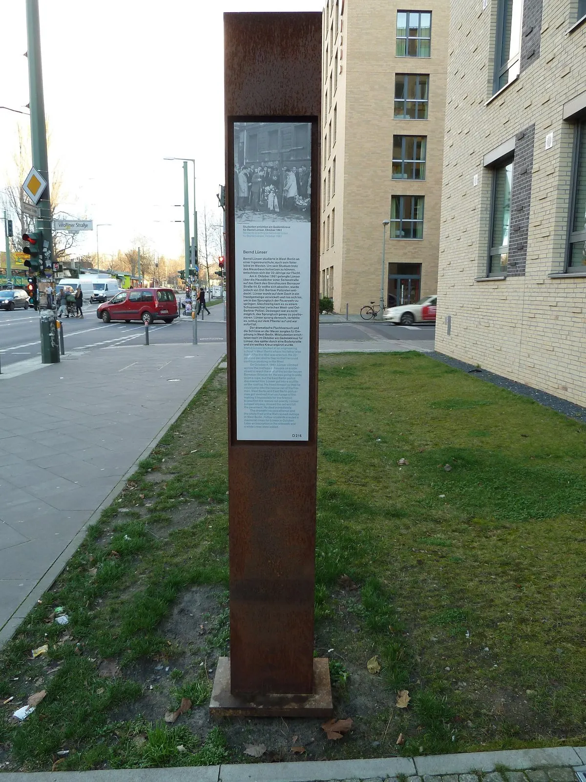 Rusted steel stele with a photo and text panel on a strip of grass beside a street and modern buildings