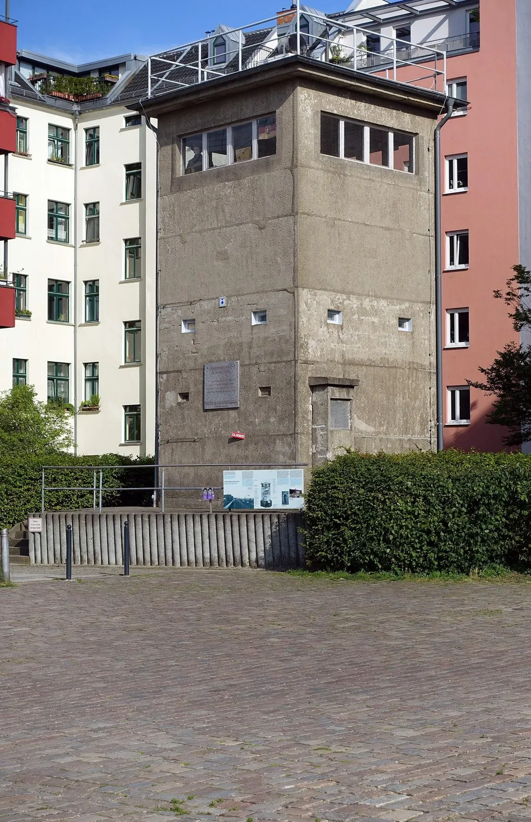 Square concrete former border watchtower among apartment buildings, with a plaque and information boards at its base