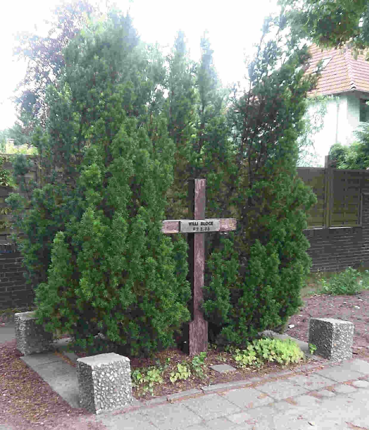 Wooden cross with a name plate standing between two tall conifers, with concrete bollards and paving in front