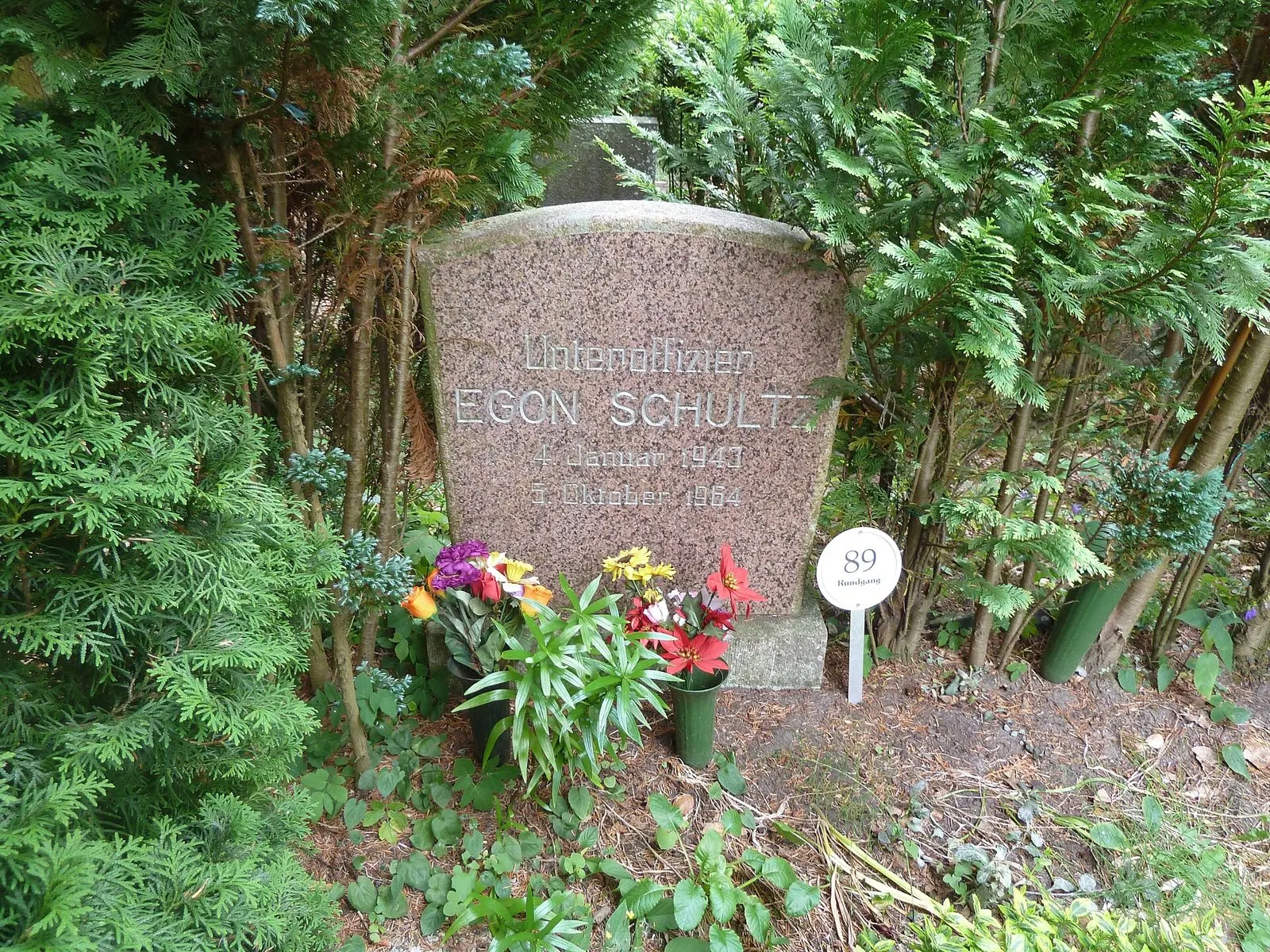 Pink granite headstone inscribed 'Unteroffizier Egon Schultz' with dates, flowers planted in front, among conifers