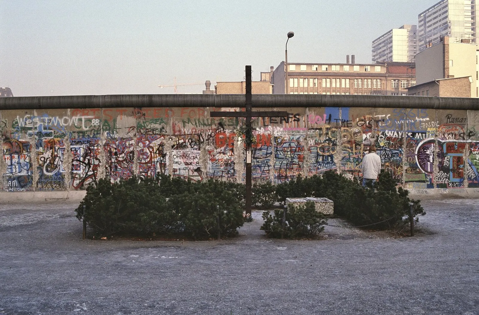Tall dark wooden cross in a planted bed before the graffiti-covered Berlin Wall