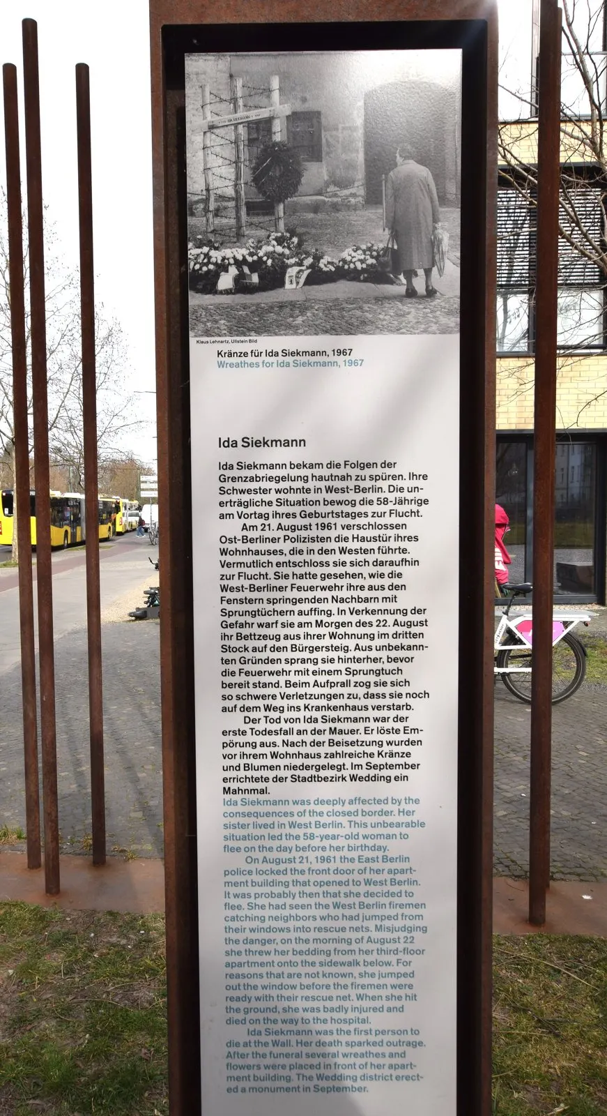 Rusted steel stele with a historical photo of a memorial with wreaths and a text panel about Ida Siekmann, among steel rods