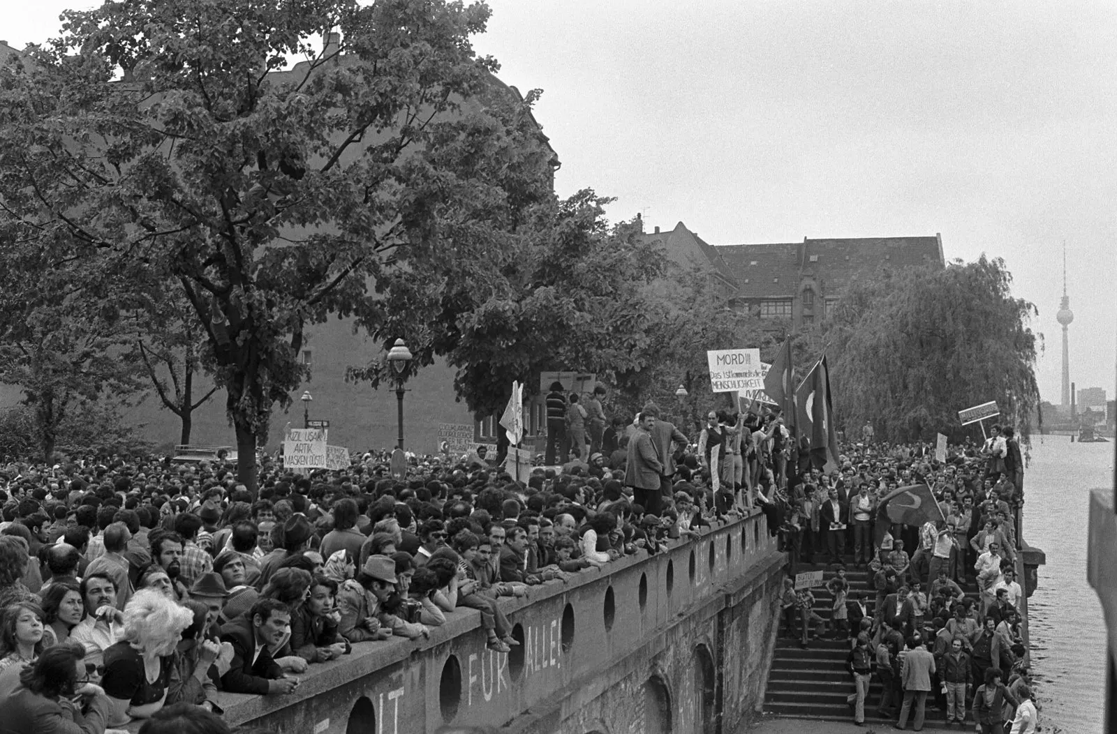 Black-and-white photograph of a large crowd gathered along a riverside wall and steps, some holding placards, with the television tower in the distance