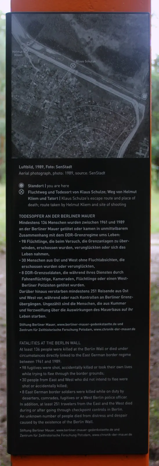 Dark information panel on an orange column with an aerial photograph of the border strip and houses, marked with circled crosses and labels