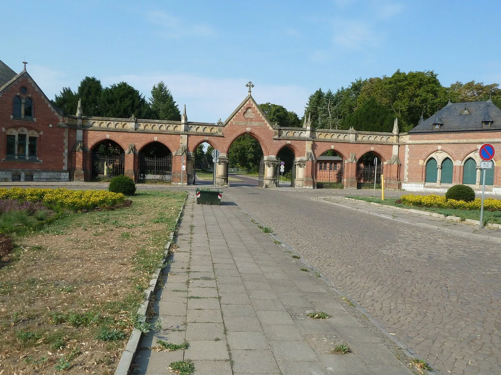 Red-brick cemetery gateway with pointed arches and a small cross on top, a paved path and flower beds in front, under a blue sky