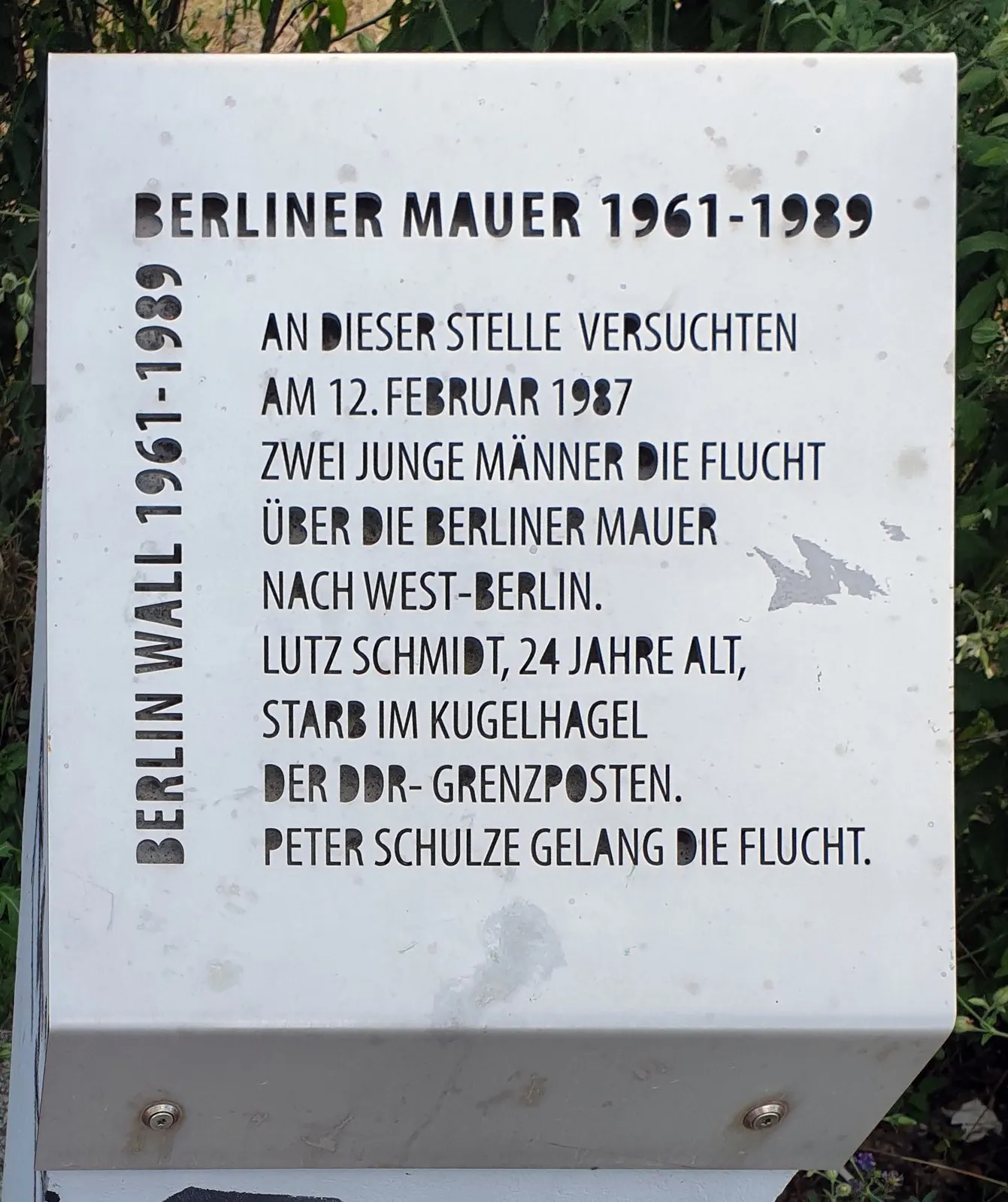 White metal plaque with cut-out lettering: "An dieser Stelle versuchten am 12. Februar 1987 zwei junge Männer die Flucht ... Lutz Schmidt, 24 Jahre alt"