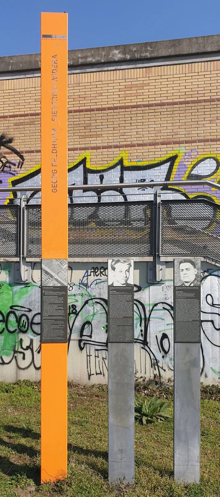 Orange post and two grey steles with portraits in front of a brick wall and railings