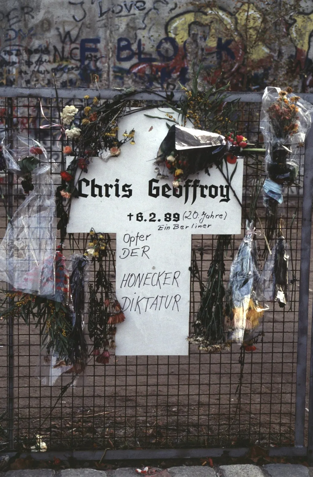 White cross on a mesh fence covered in flowers, hand-lettered with his name, a date, his age and "Opfer der Honecker Diktatur", with the graffiti-covered Wall behind