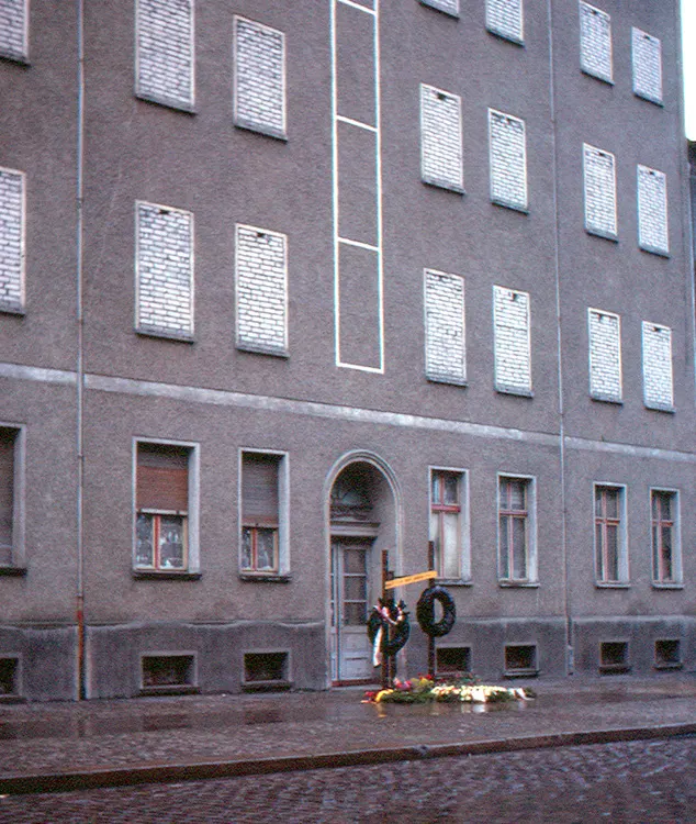 Grey apartment building with bricked-up windows; at the door a cross, wreaths and flowers on the pavement