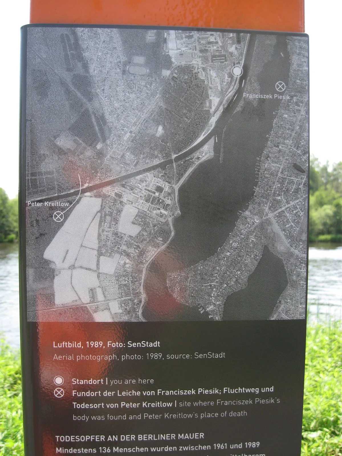 Panel with a black-and-white aerial photograph of a river and lake with labelled marks, above German text
