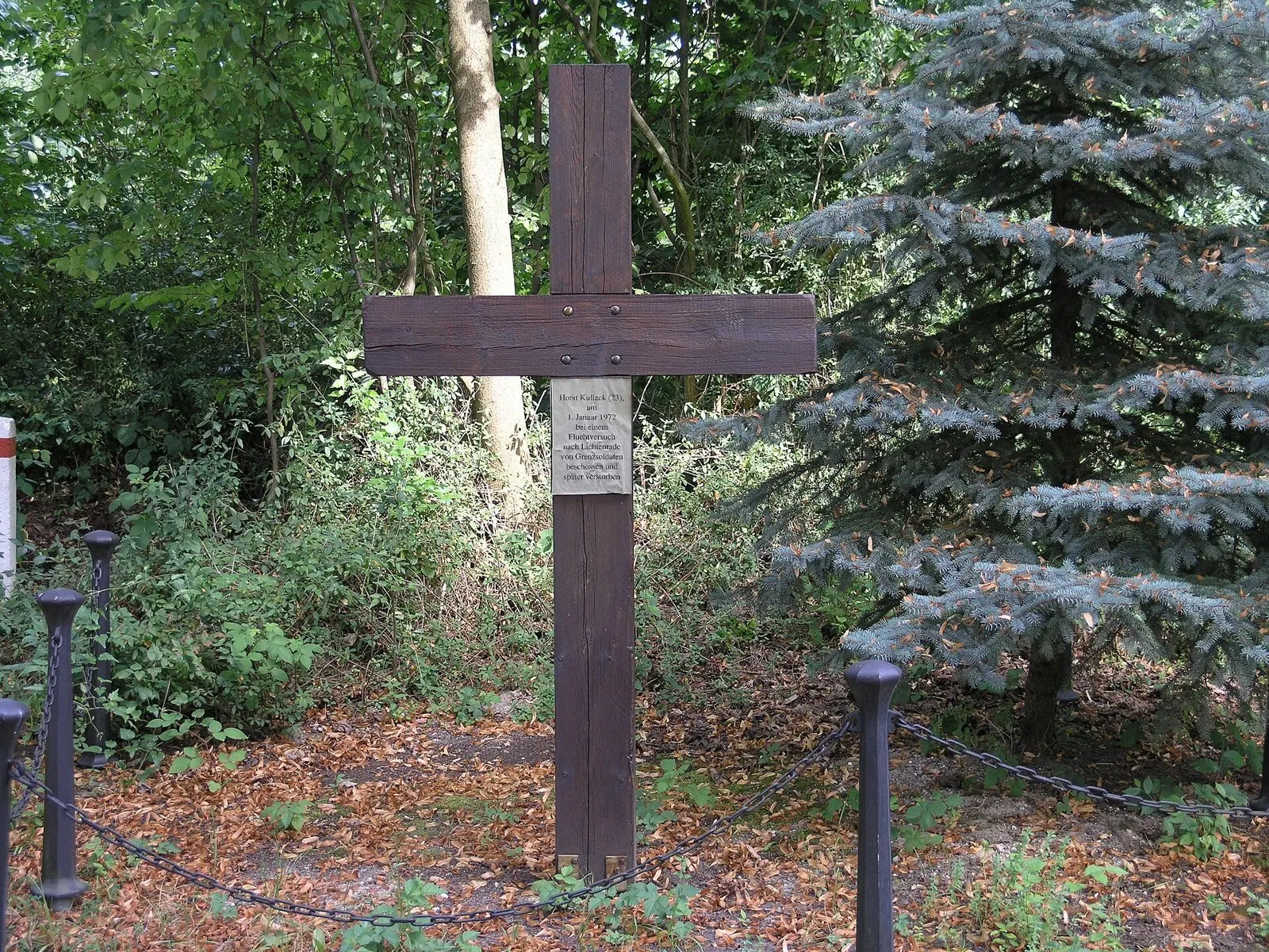 Tall dark wooden cross with a small plaque, surrounded by a low chain fence among trees