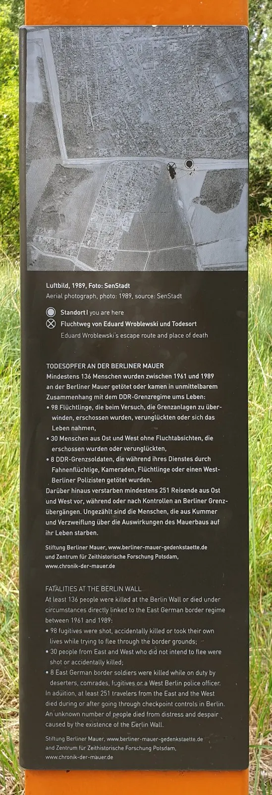 Panel with a black-and-white aerial photograph of fields and settlements with a marked point, above German and English text