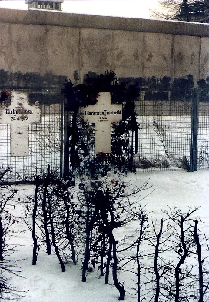 Snowy scene with a white cross lettered with her name, "18 Jahre" and a date, hung on a fence in front of the Wall, with another cross to the left