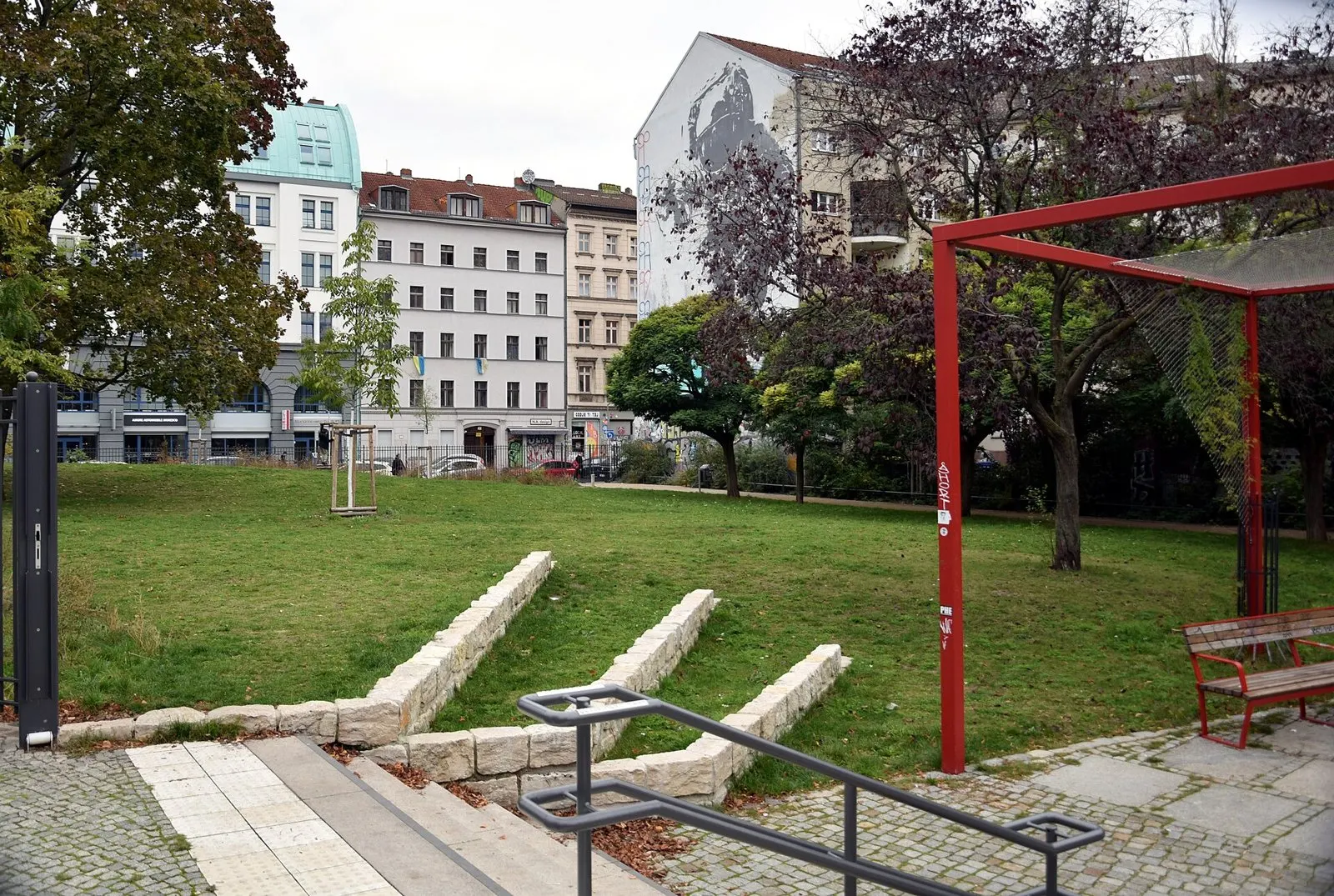Small urban park with a lawn, low stone steps, trees and a red metal frame, surrounded by apartment buildings