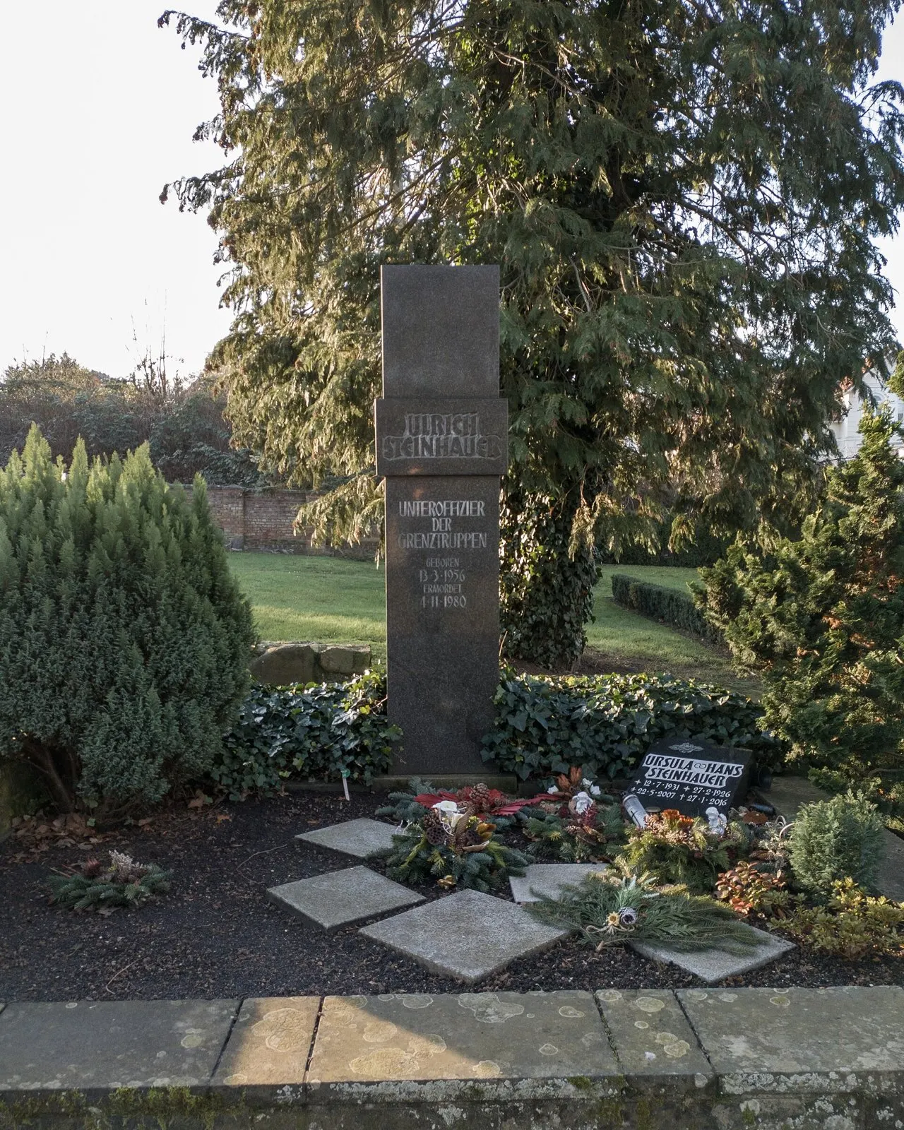 Tall dark stone with his name and inscription in a grave bed with wreaths and evergreens, under a large conifer