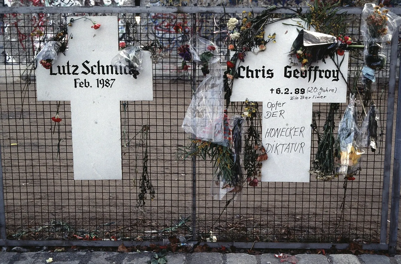 Two white crosses on a mesh fence, hung with flowers; the left one lettered "Lutz Schmidt Feb. 1987", the right one with Chris Gueffroy's name (misspelled) and a date