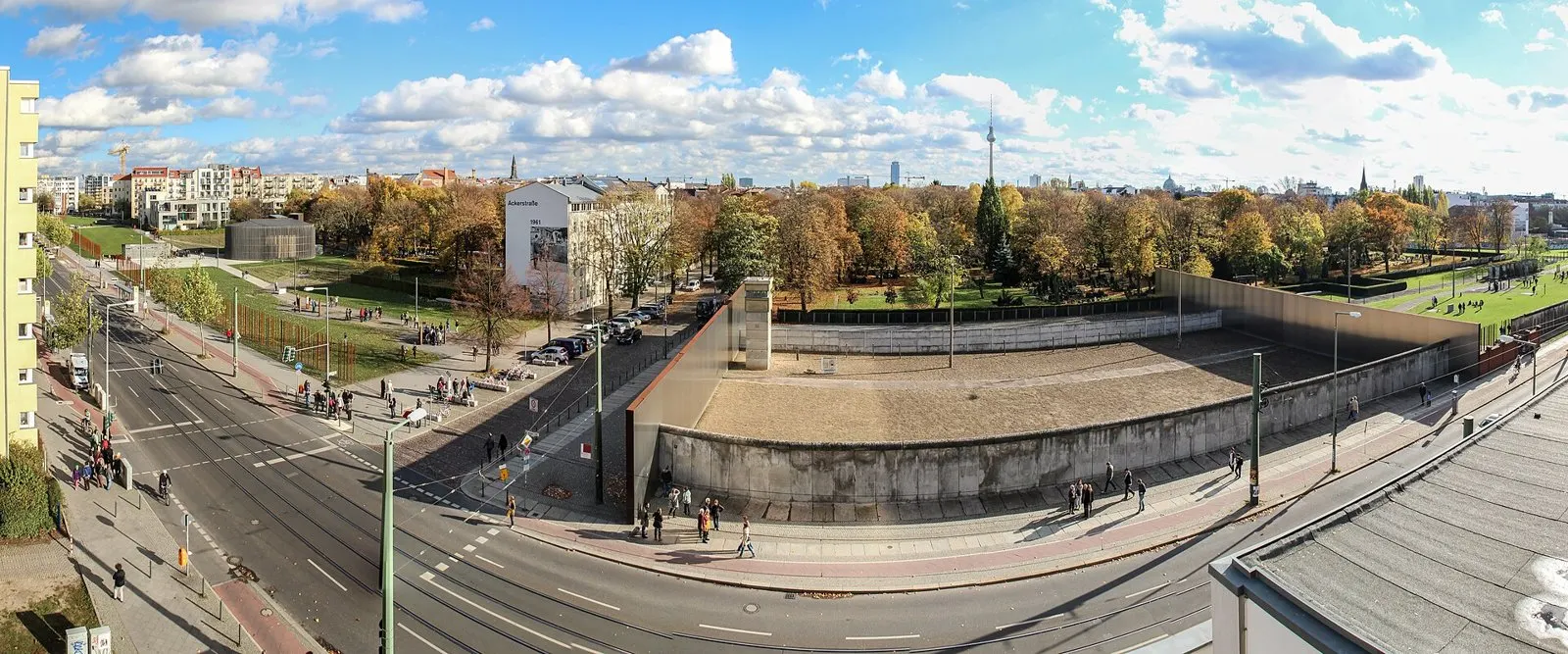 Wide elevated panorama of Bernauer Straße: a preserved section of the Wall enclosing a border strip with a watchtower, visitors on the pavement, autumn trees and the Berlin TV tower on the skyline under a blue sky with clouds.
