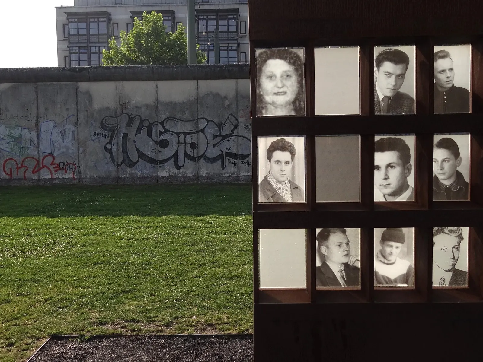 Close view of lit niches in a rusted steel frame holding black-and-white portrait photographs of men and a woman, some niches empty; beyond, a lawn and a grey section of the Berlin Wall with graffiti.