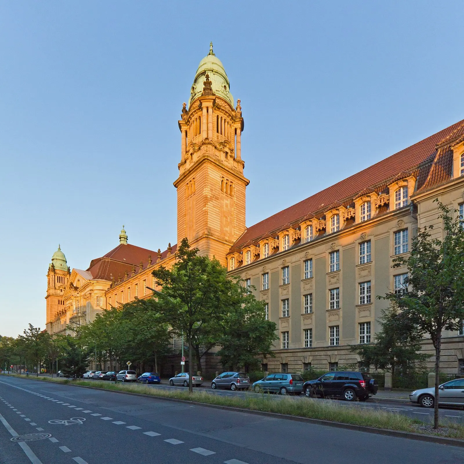 A long sandstone court building with a tall tower topped by a green copper dome, lit by low evening sun, along a wide street with parked cars and young trees.