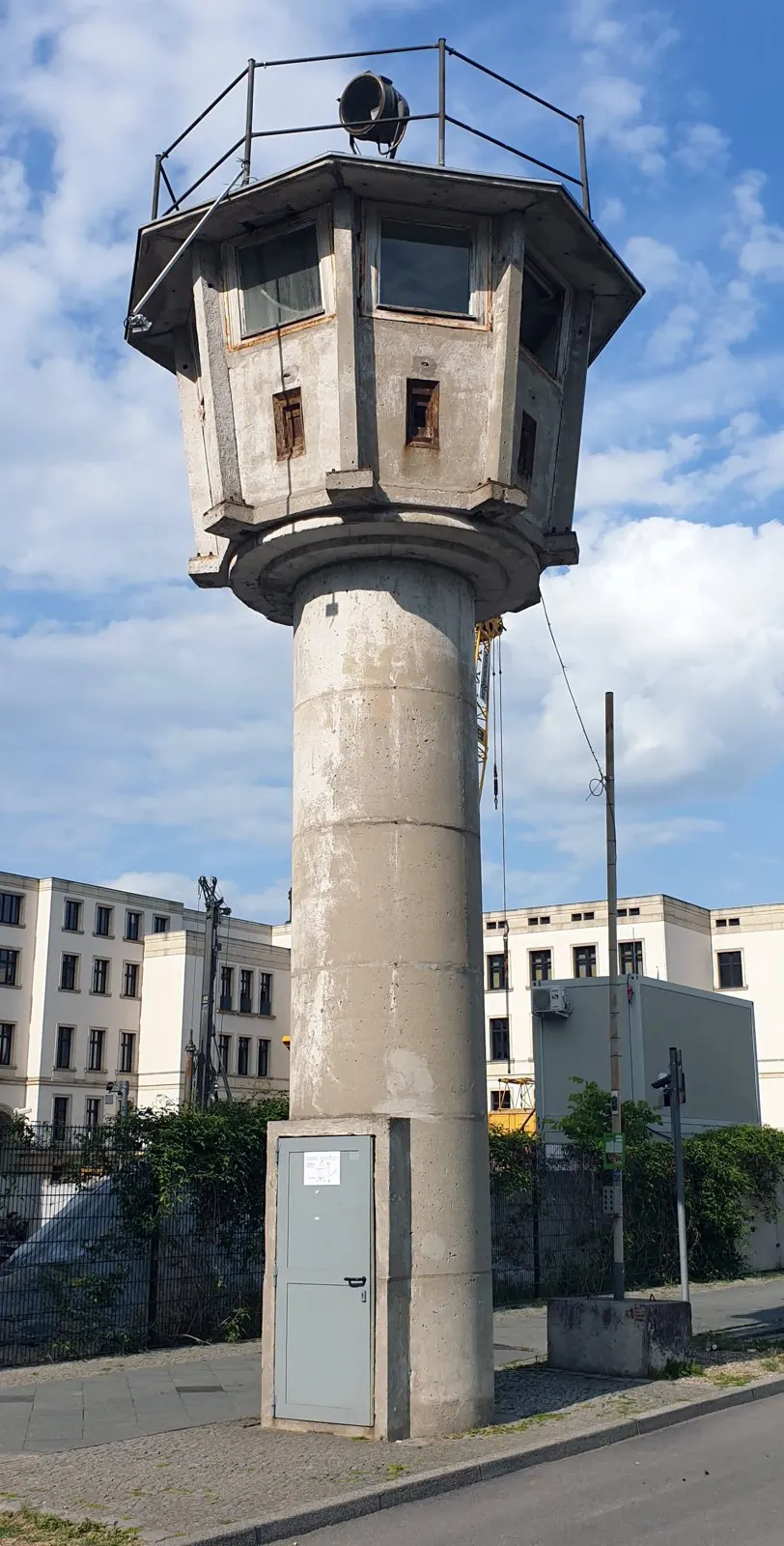 A slender round concrete watchtower with an octagonal observation cabin, a searchlight and a railing on top stands on a pavement against a blue sky with white clouds; office buildings behind.