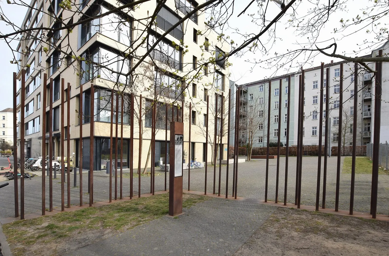 Rows of tall rusted steel rods and a rust-coloured information stele stand on a paved square in front of modern apartment buildings, seen through bare branches.