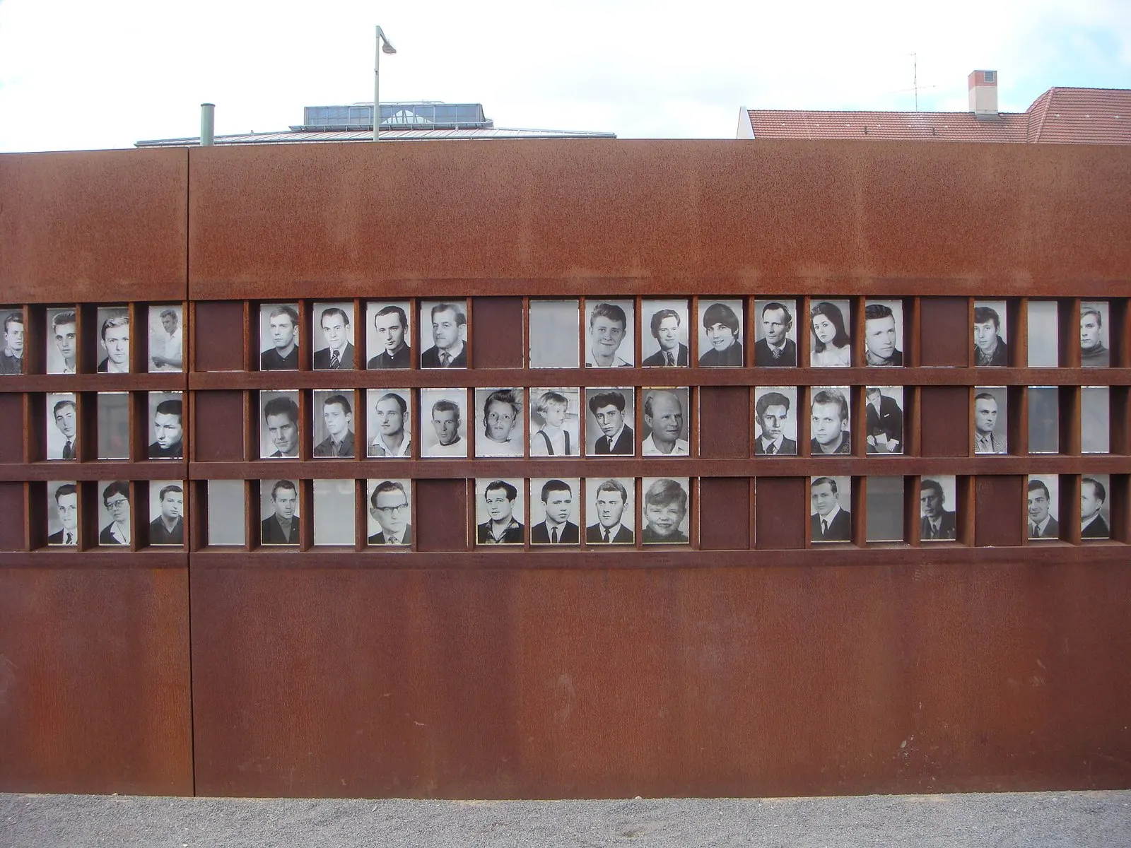 Frontal view of a rust-coloured steel wall with three rows of small black-and-white portrait photographs, several windows left blank; a street lamp and rooftops rise behind it under a white sky.