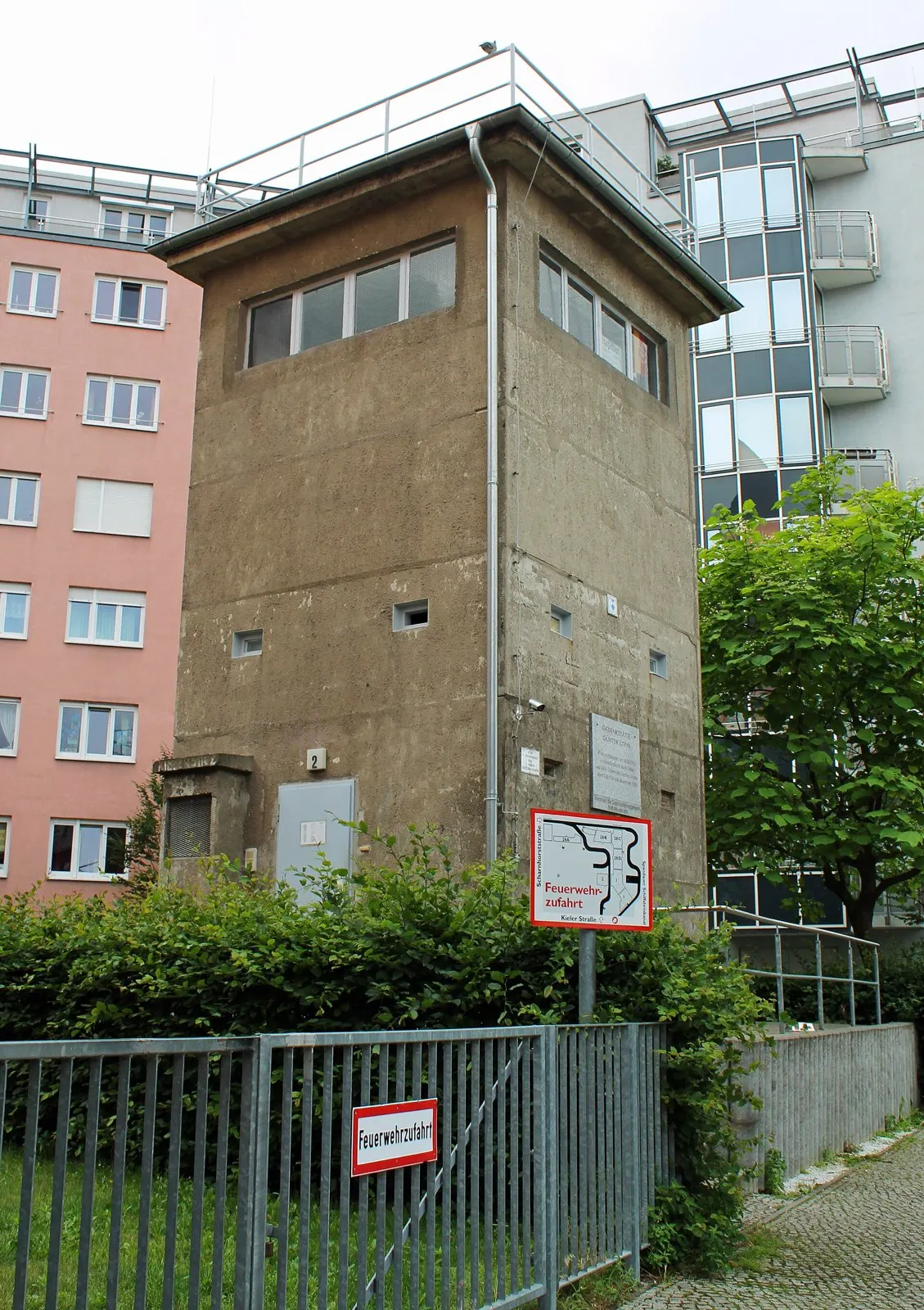 A square, brownish concrete watchtower with a band of windows and a railing on its flat roof stands behind a metal fence with a fire-lane sign, between pink and glass-fronted apartment buildings.