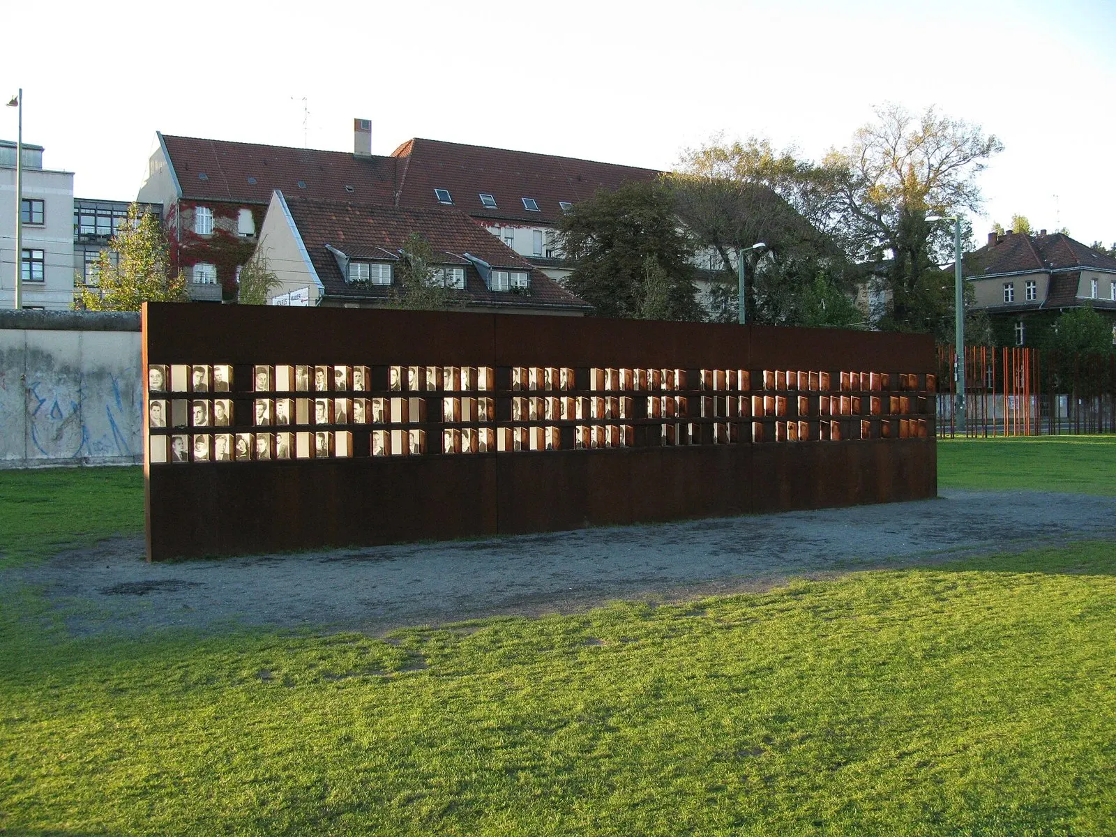 A long rust-coloured steel wall with rows of small black-and-white portrait photographs stands on a lawn in warm, low sunlight; a grey section of the Berlin Wall is on the left and rusted steel rods on the right, with red-roofed houses behind.