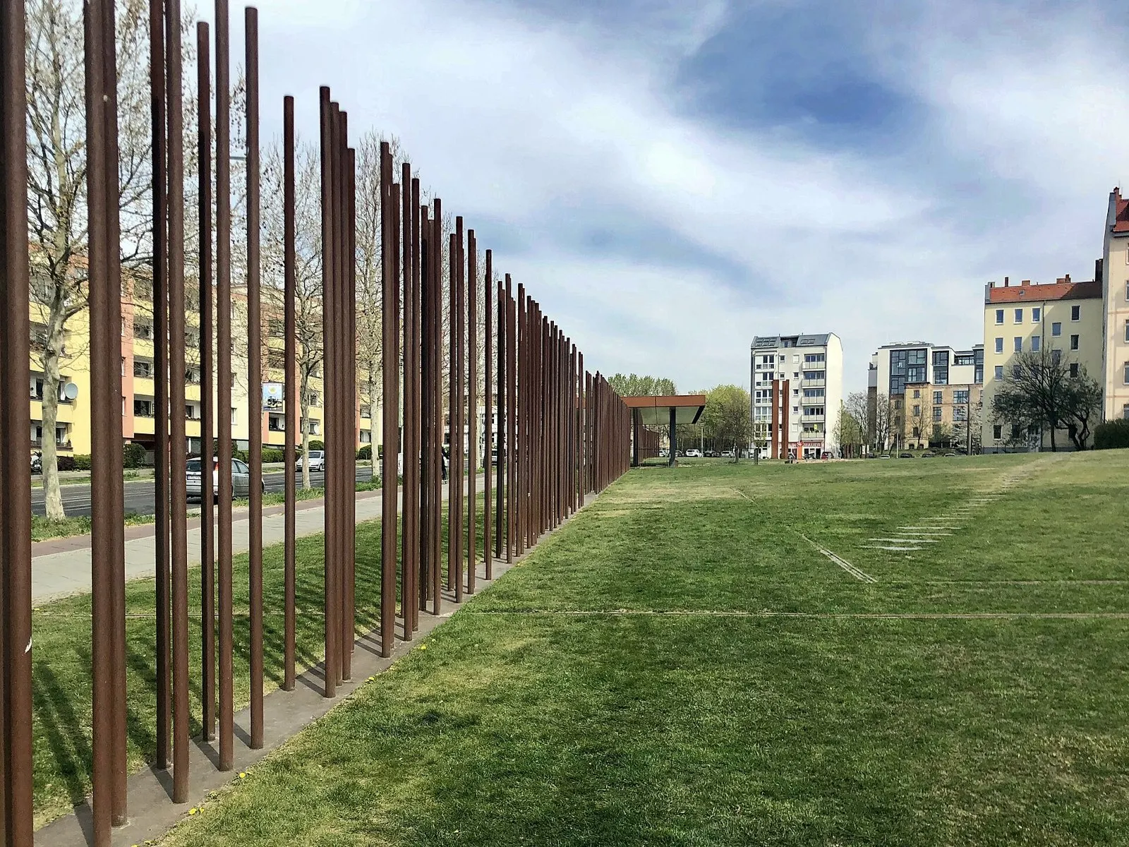A long line of tall rusted steel rods stands along the edge of a lawn, receding into the distance under a cloudy blue sky, with apartment buildings on both sides.