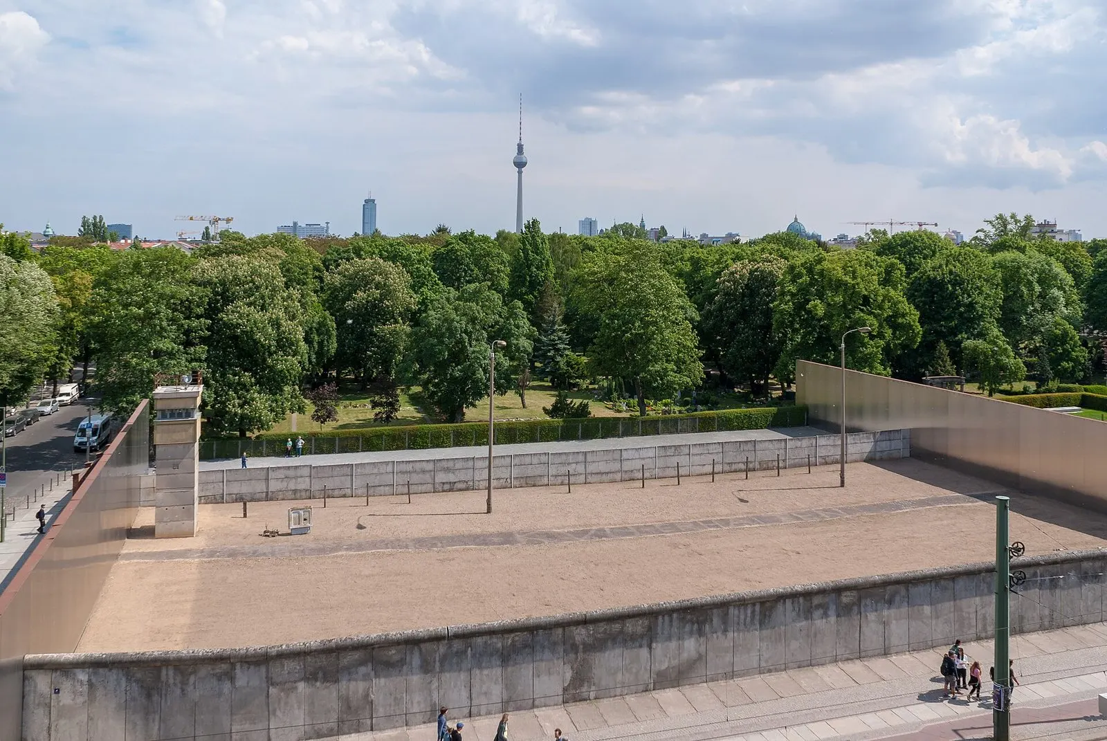 Elevated view over a preserved section of the former border strip: a sandy strip with lamp posts and a watchtower between two concrete walls, trees beyond and the Berlin TV tower on the skyline.