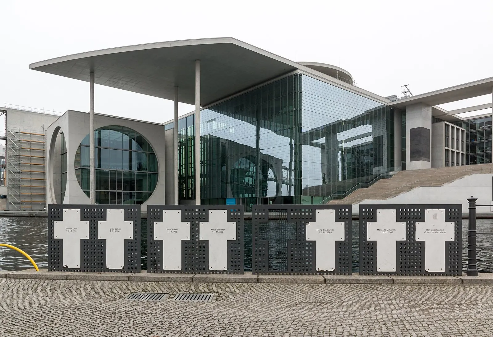 Seven white crosses with names and dates on dark perforated metal panels stand along a river railing; behind them are the water and a large modern glass-and-concrete building.