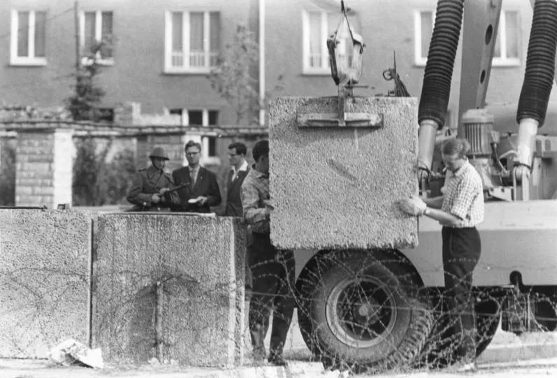 Black-and-white photograph: a crane lowers a large concrete block beside other blocks behind barbed wire; a worker guides it while a man in uniform with a gun and men in suits watch in front of a building.