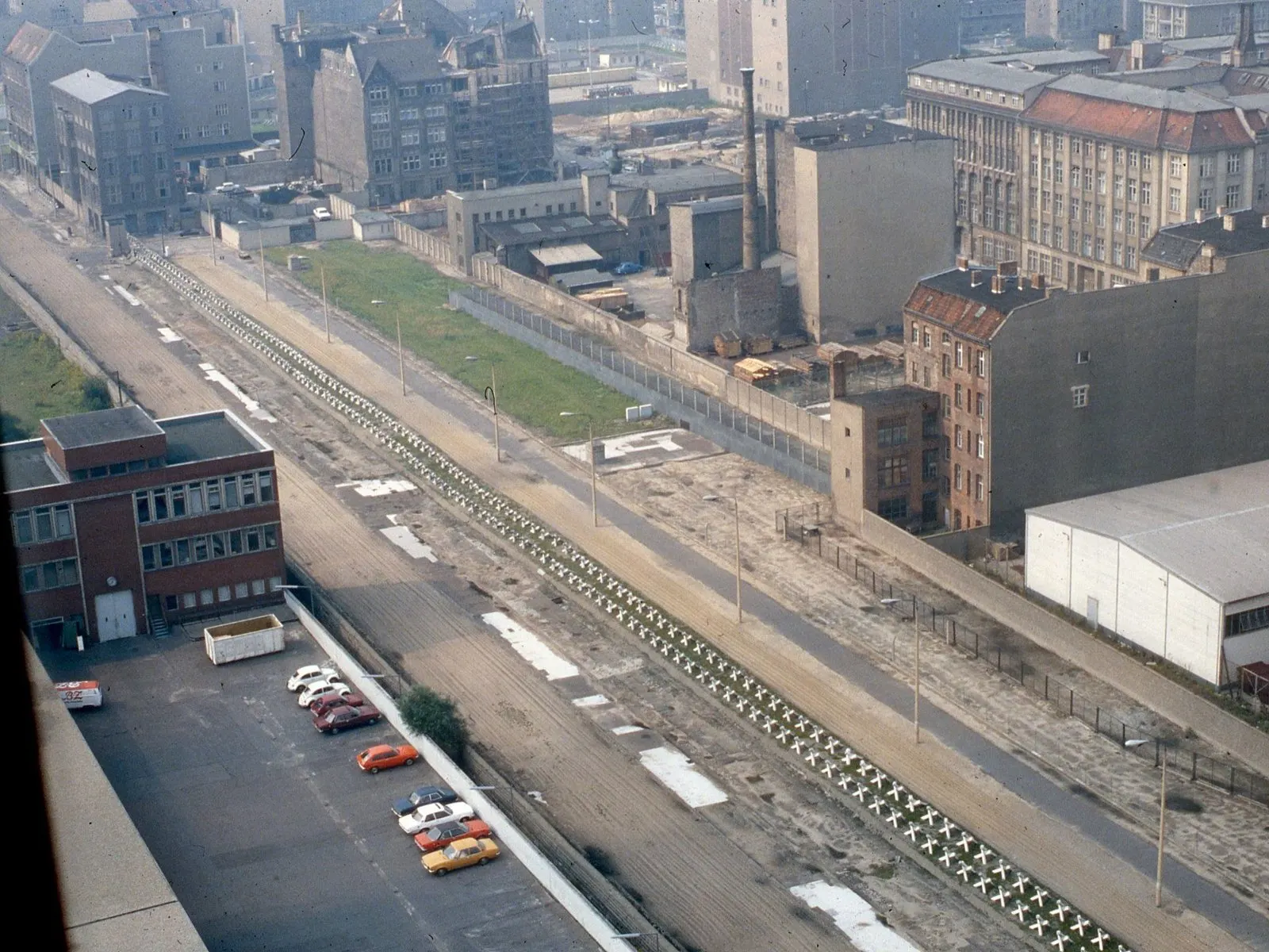 Elevated colour photograph of the border strip: a wide sandy strip with a long line of anti-vehicle obstacles and a patrol road runs between walls, lined by old apartment and factory buildings; cars are parked in the foreground.